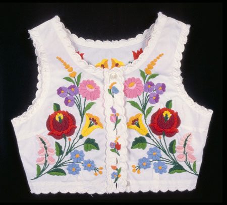 Sleeveless white cotton vest with multi-coloured embroidered floral motif. It has 5 buttons and a zigzag trim., © CMC/MCC, 76-514.2