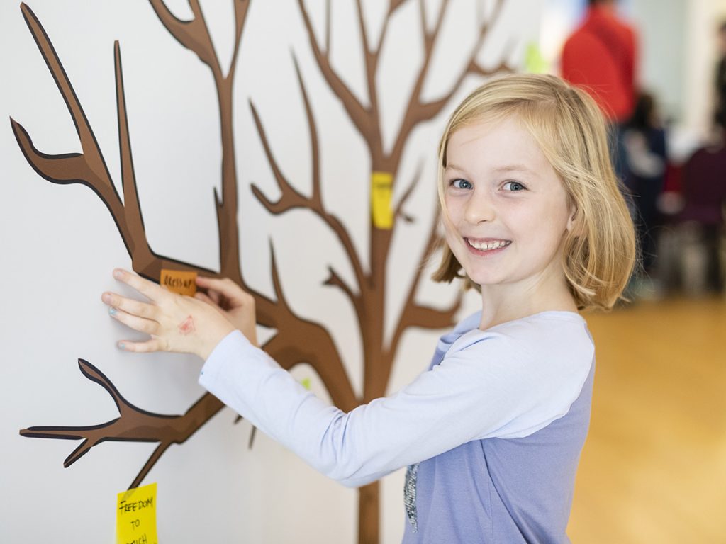 Young girl in a blue shirt attaching a sticker to an image of a tree on a wall