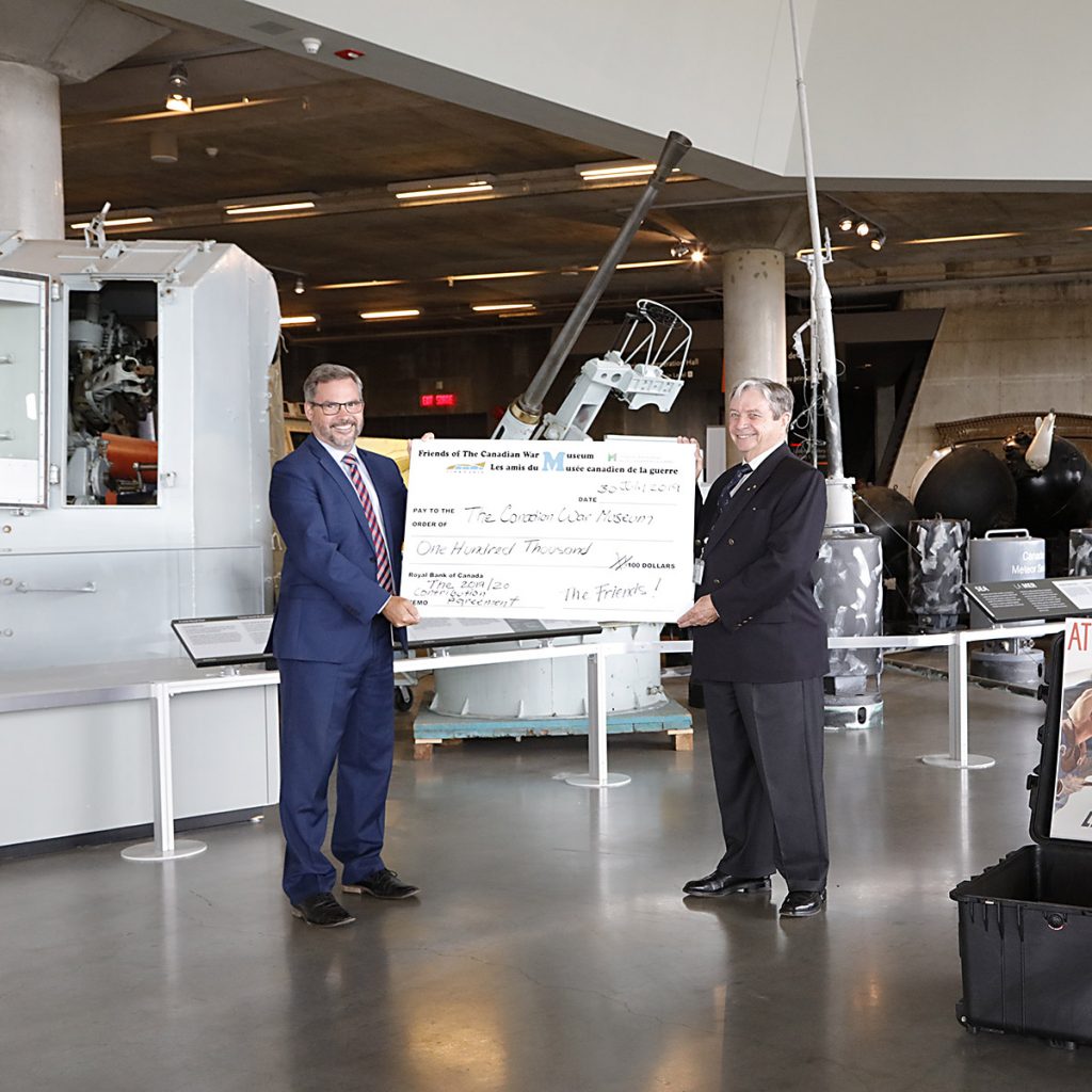 Two smiling men stand holding a large cheque, surrounded by military vehicles