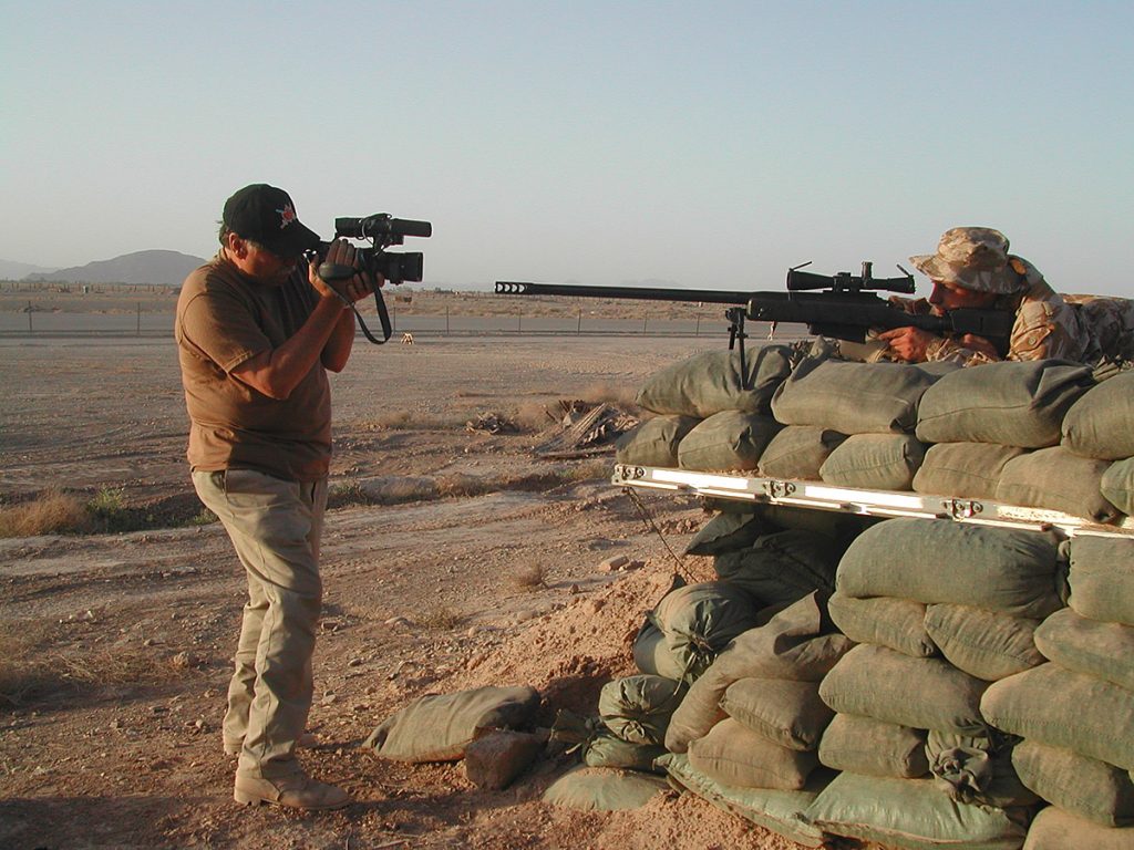 A man filming a soldier who is aiming a gun while standing behind a pile of sandbags in a desert setting
