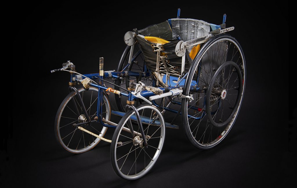 A wheelchair with a blue metal frame, four wheels and a bucket seat covered in silver duct tape