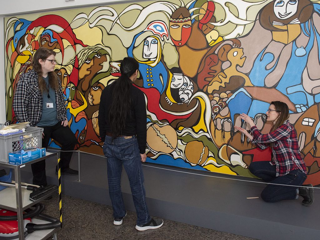 A crouching woman speaks to two standing men while gesturing to a colourful painted mural