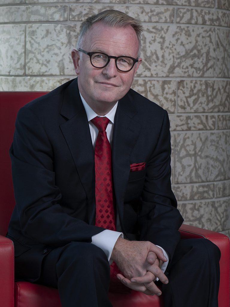 Portrait of a smiling man seated in a red armchair, wearing a dark suit, white shirt, red tie and glasses