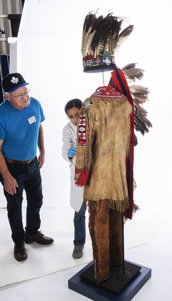 A man in jeans, t-shirt and baseball cap, and a woman in a white lab coat, examine Indigenous regalia displayed on a mannequin