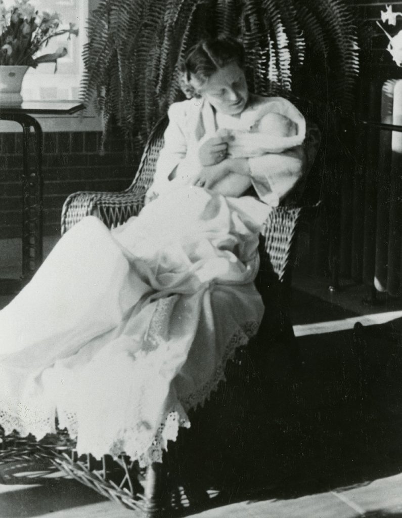 A black-and-white image of a woman in a long white robe holding a baby, while seated in a woven wicker chair