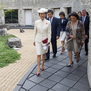Group of people walking outdoors on a stone walkway bordered by gravel and plants, led by a woman in a white dress suit and hat