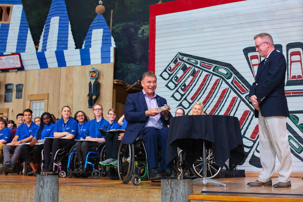 Hansen in a wheelchair holding his glove, on the Grand Hall stage with students and Mark O’Neill