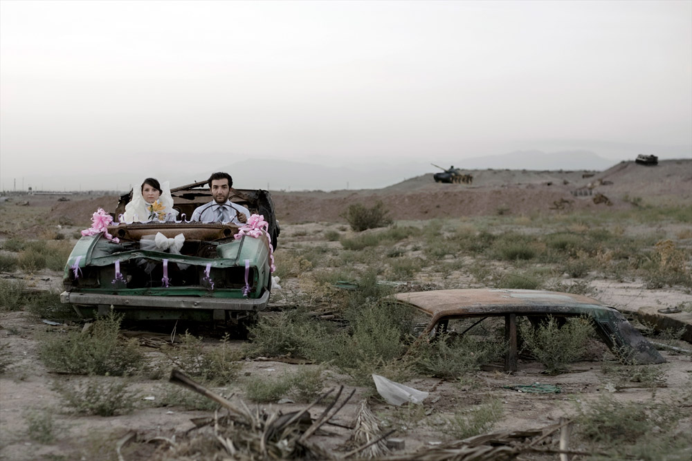 A newly married couple sits in an old car, in a war-torn landscape
