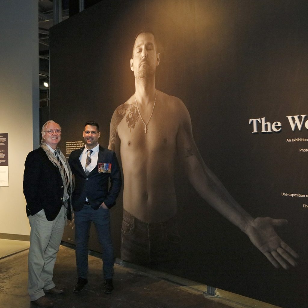 Two men posing in front of a large photograph of a shirtless man with tattoos and a long scar on his arm