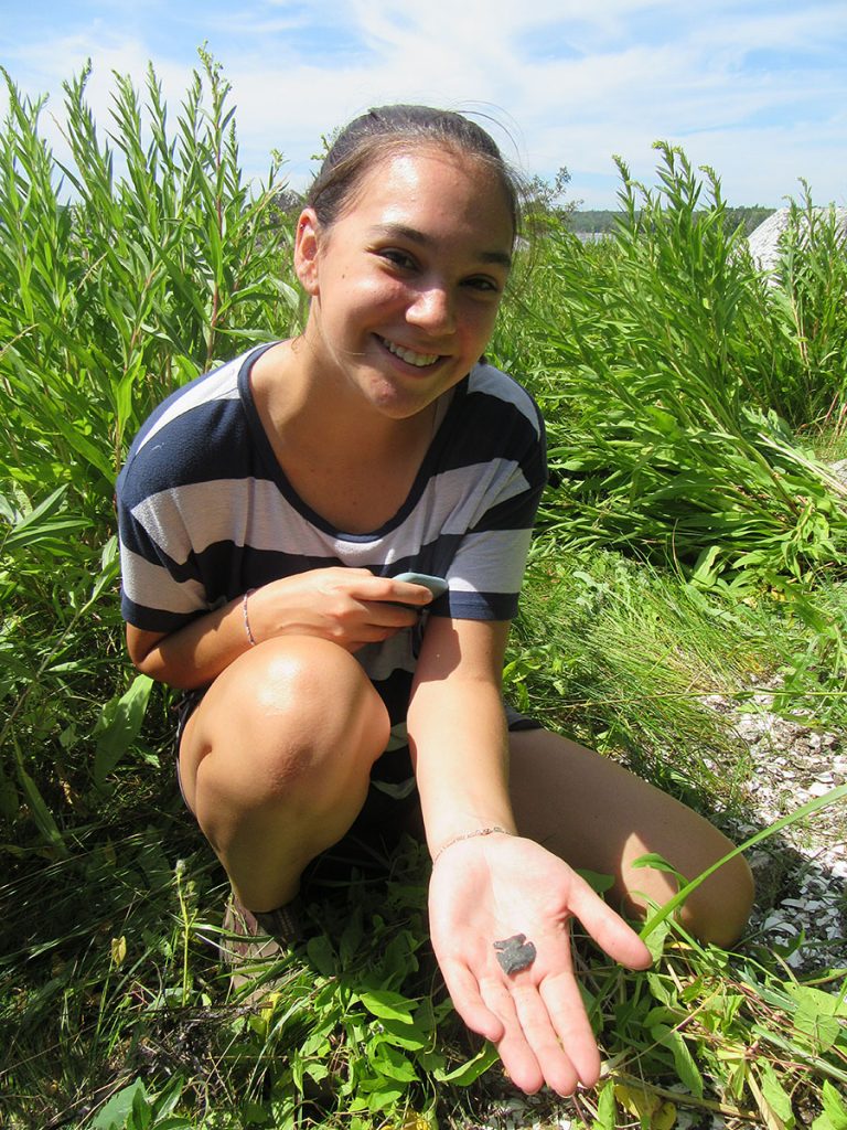 A young, smiling woman crouches in green grass and displays a stone arrowhead in the palm of her hand