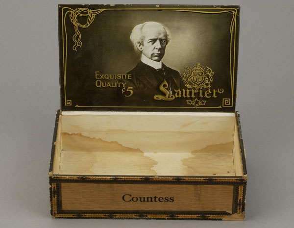 Early rectangular wooden cigar box with hinged lid. The top of the lid is etched with the word "Laurier" inside a wreath. The inside has a black-and-white photograph of Laurier with a crest stamped in gold. Canadian Museum of History, 1999.124.21