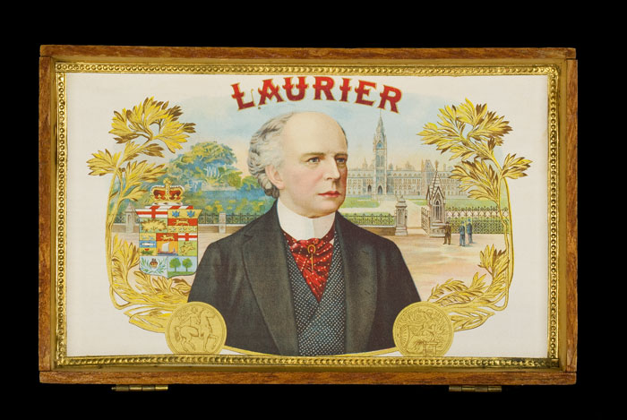 Early rectangular wooden cigar box with hinged lid. The middle of the outer lid features a painting of Laurier’s head, encircled by a metal wreath. The inner lid has a full-colour image of Laurier, depicted against the original Parliament Buildings in Ottawa. Canadian Museum of History, 1999.124.18