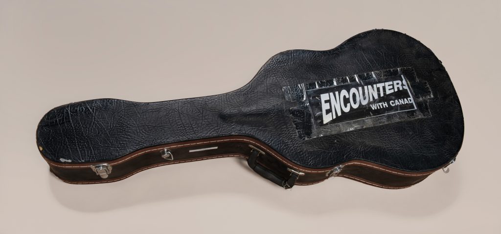 A black leather guitar case with a large black and white sign taped on, reading “Encounters with Canada.”