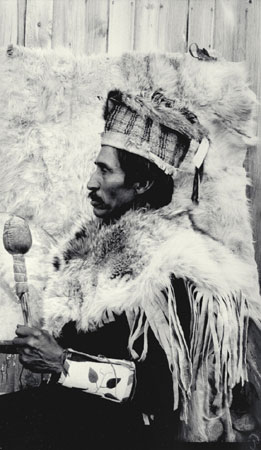 Ususéllst, a Nlaka'pamux (Thompson) man wearing traditional clothing (fringed fur cape, woven bark and fur hat, beaded cuffs and rattle) , profile view, photographed in 1913 at Spences Bridge, British Columbia, © CMC/MCC, J.A. Teit, 20831
