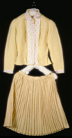 Part of a costume that was made for children to wear during dances and plays, this commercially made blouse is made with yellow synthetic cloth, long sleeves, and decorated with white lace and floral ribbons., © CMC/MCC, 77-251.1
