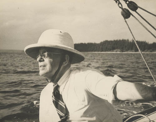 A man in a pith helmet and glasses and tie sailing a boat, with water and trees in the background.