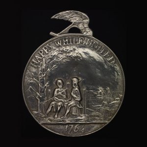 Circular medal featuring two figures sitting side by side, with the words “Happy while united” and the year 1764.