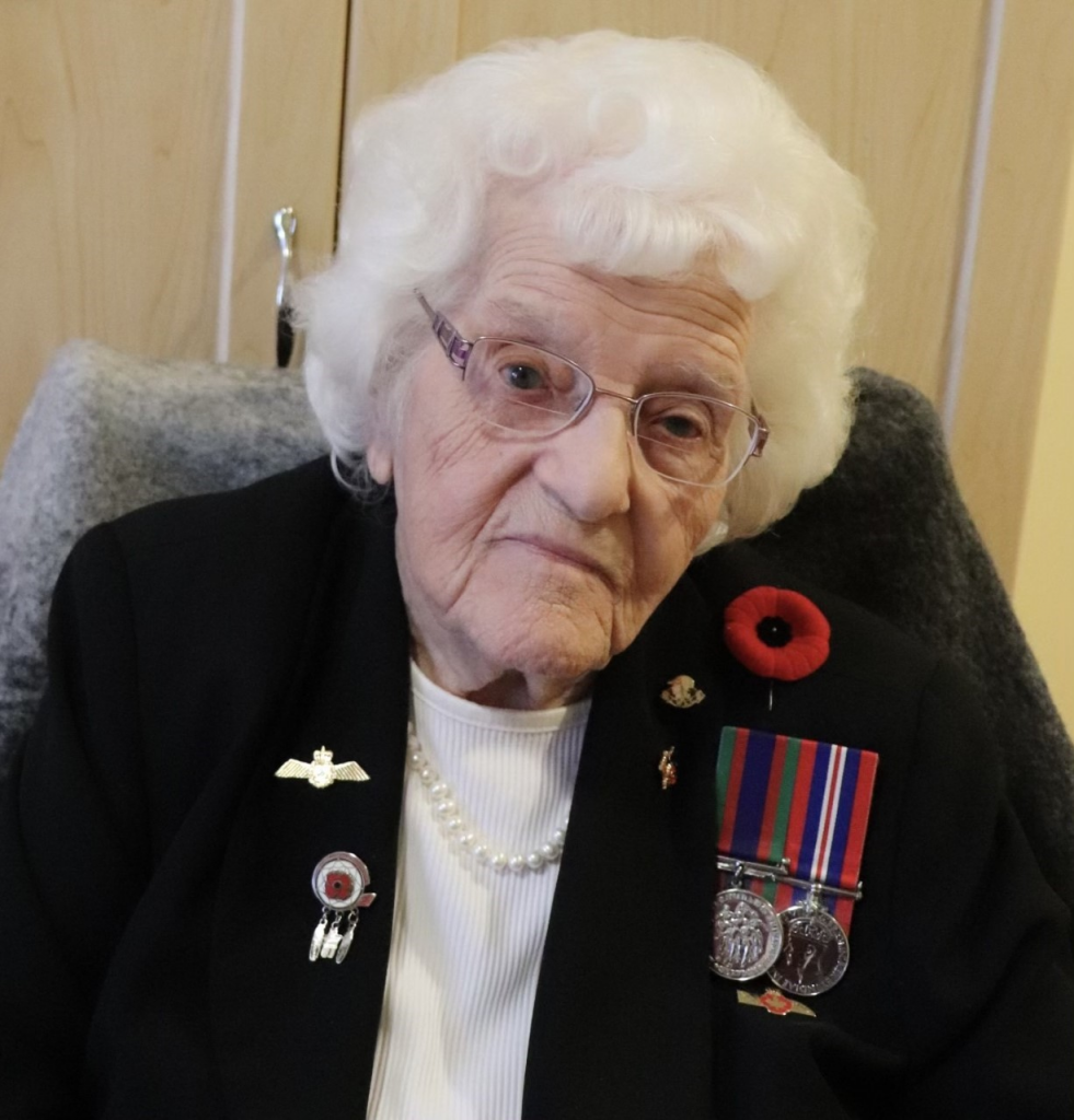 A woman with white hair sits, wearing a poppy, pins, and military medals.