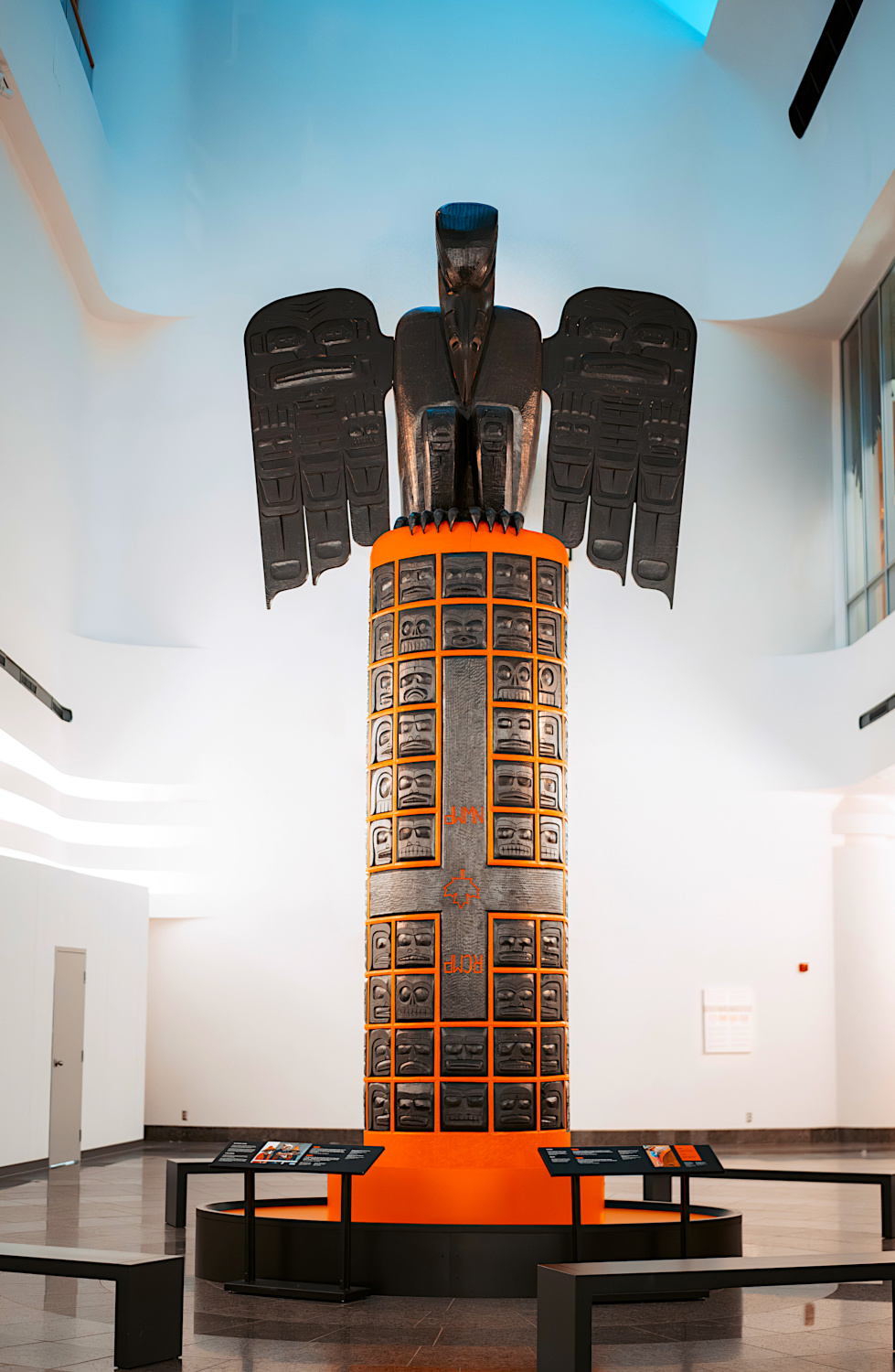 A large cylindrical sculpture featuring children’s faces painted black within an orange grid. A large carved black raven looks down upon the children from the top.