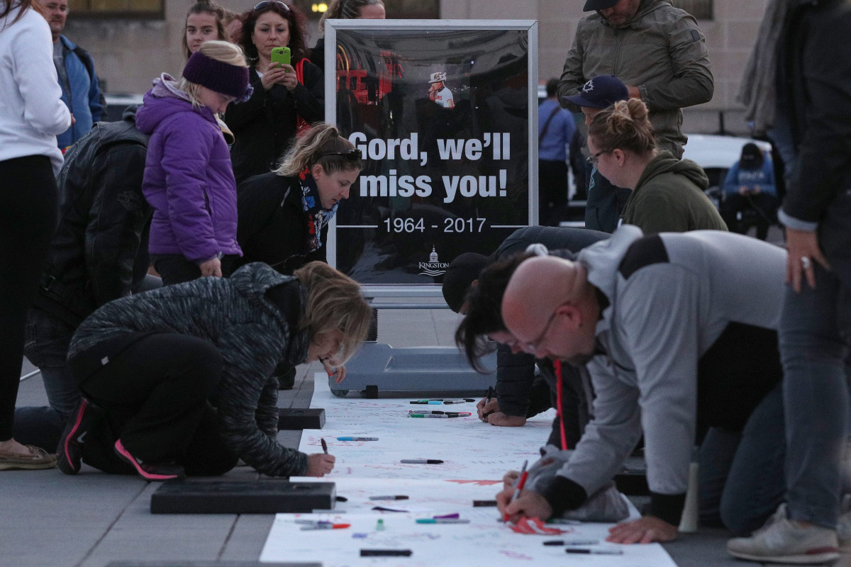 Several adults kneel on the ground, writing on a large piece of paper with markers. More adults and children stand nearby and a large sign in the background reads “Gord, we’ll miss you! 1964-2017.”