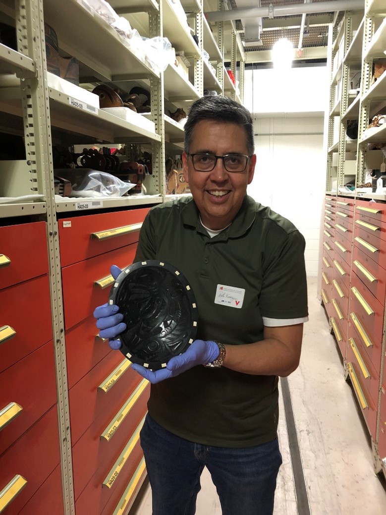 An Indigenous man with short dark hair, glasses, and blue gloves holds a black, carved stone bowl, while standing in an aisle between floor-to-ceiling storage shelves and drawers.