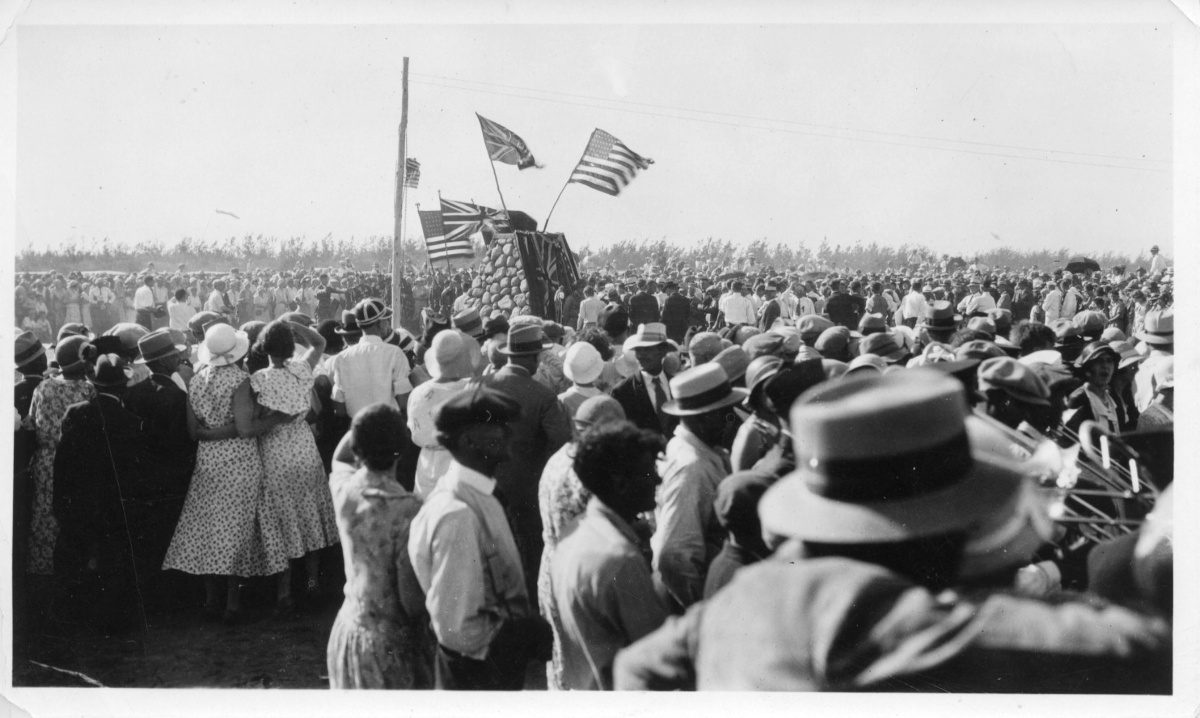 A black-and-white photo of a large crowd surrounding a stone cairn flying the Union Jack and the flag of the United States.