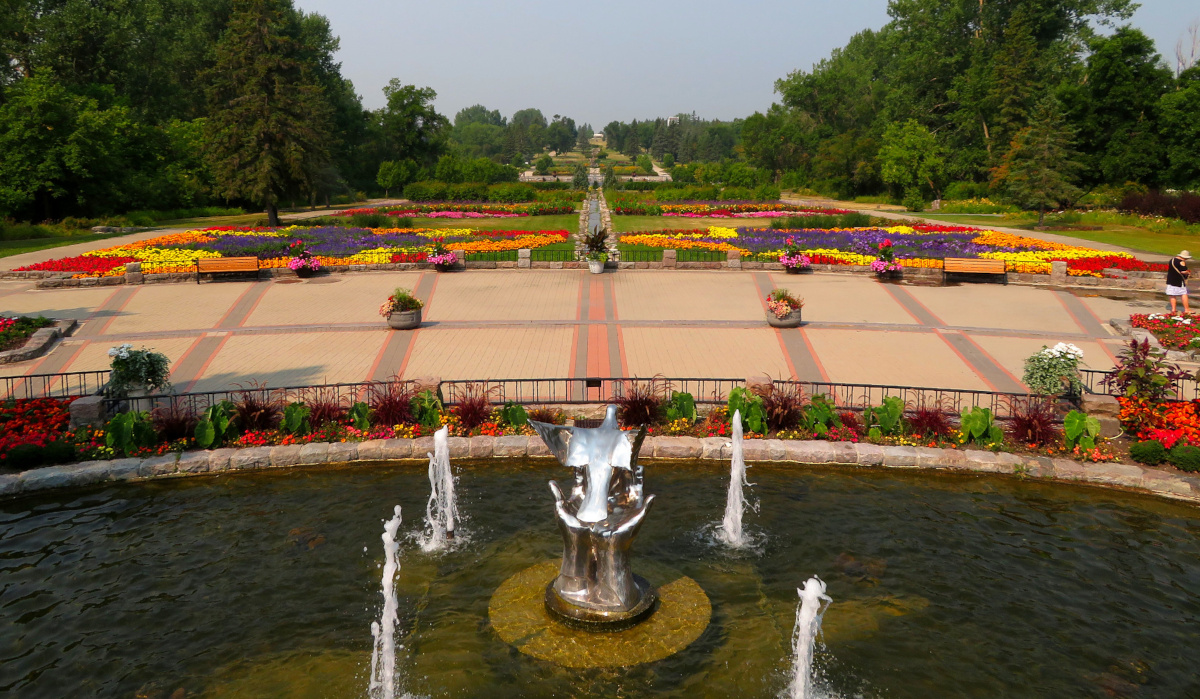 An extensive, landscaped flower garden with a pool of water and a fountain in the foreground. In the centre of the pool is a shiny metal sculpture of a pair of hands releasing a dove.