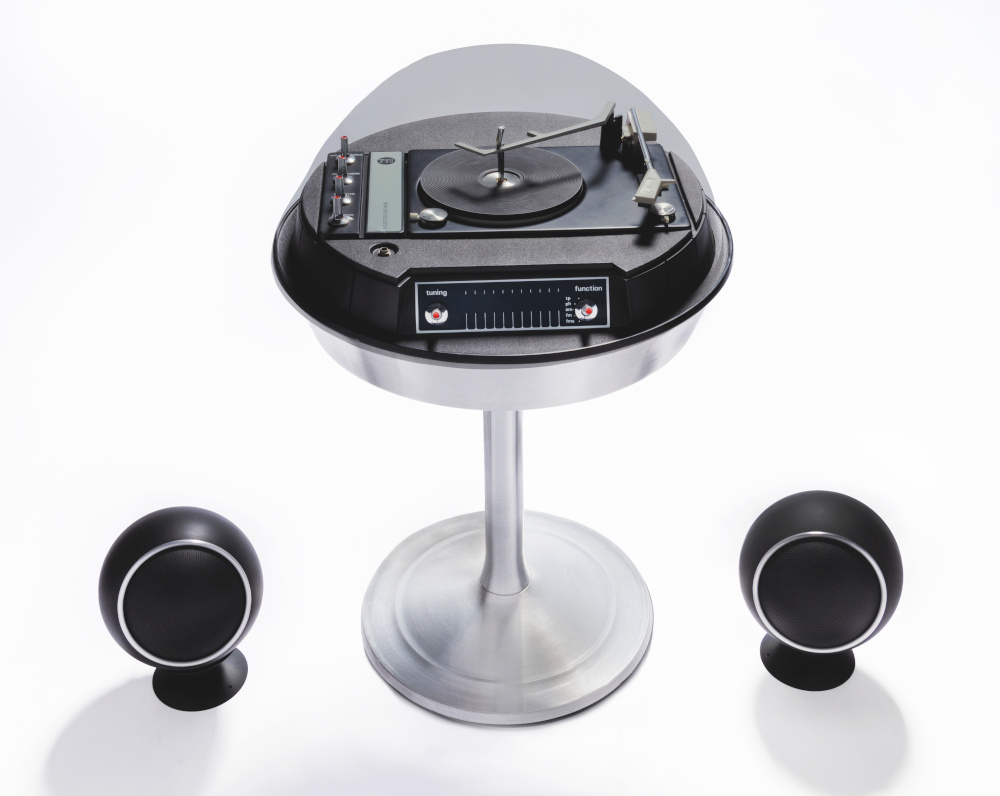 A record player with a space-age design, placed on a narrow pedestal and covered with a plastic dome, with small spherical speakers on the floor to either side.