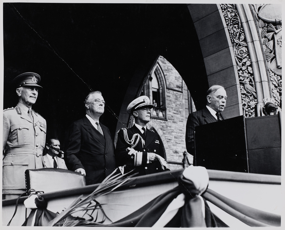 Three light-skinned men — two in military uniform, and one in a suit — stand next to a light-skinned man in a suit, who is speaking into a microphone at a podium.
