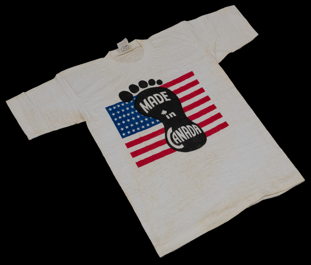 White t-shirt with red, white and blue U.S. flag, overlaid with a black footprint and white text that reads, “Made in Canada.”