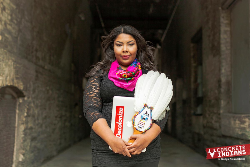 An Indigenous person with long hair, wearing a black lace dress and a pink floral scarf, holding a beaded white ceremonial feather fan and a laptop with a “Decolonize” sticker.