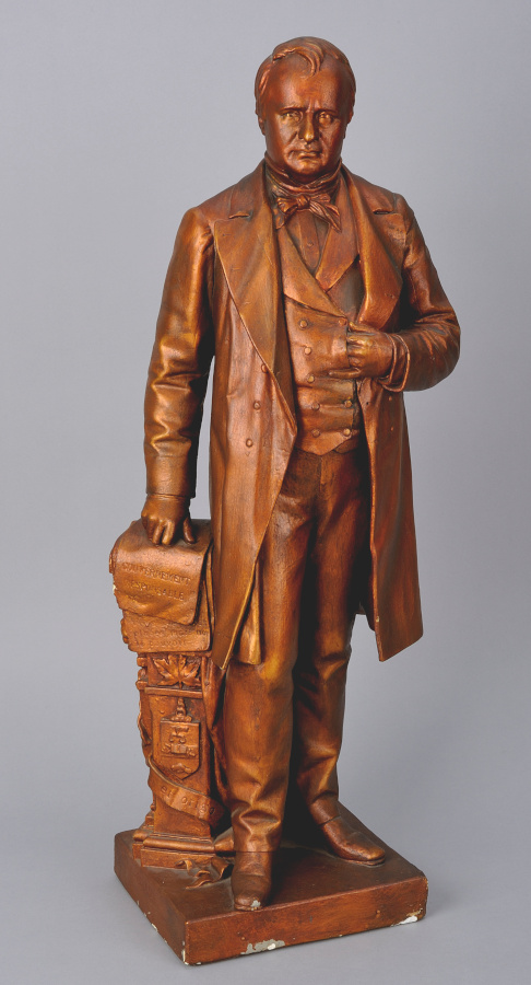 A wooden statue of a man in formal dress, leaning on a stack of books with one hand, the other hand inserted into his vest.