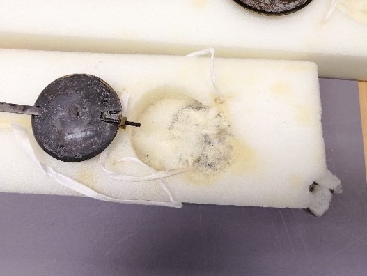 A circular metal item with a bumpy texture atop a white foam block with a matching circular indentation that is visibly stained.