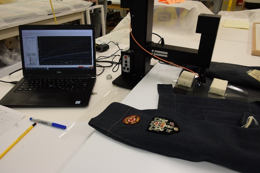 A dark jacket with military insignia positioned below the protruding arm of a black apparatus hooked up to a laptop.