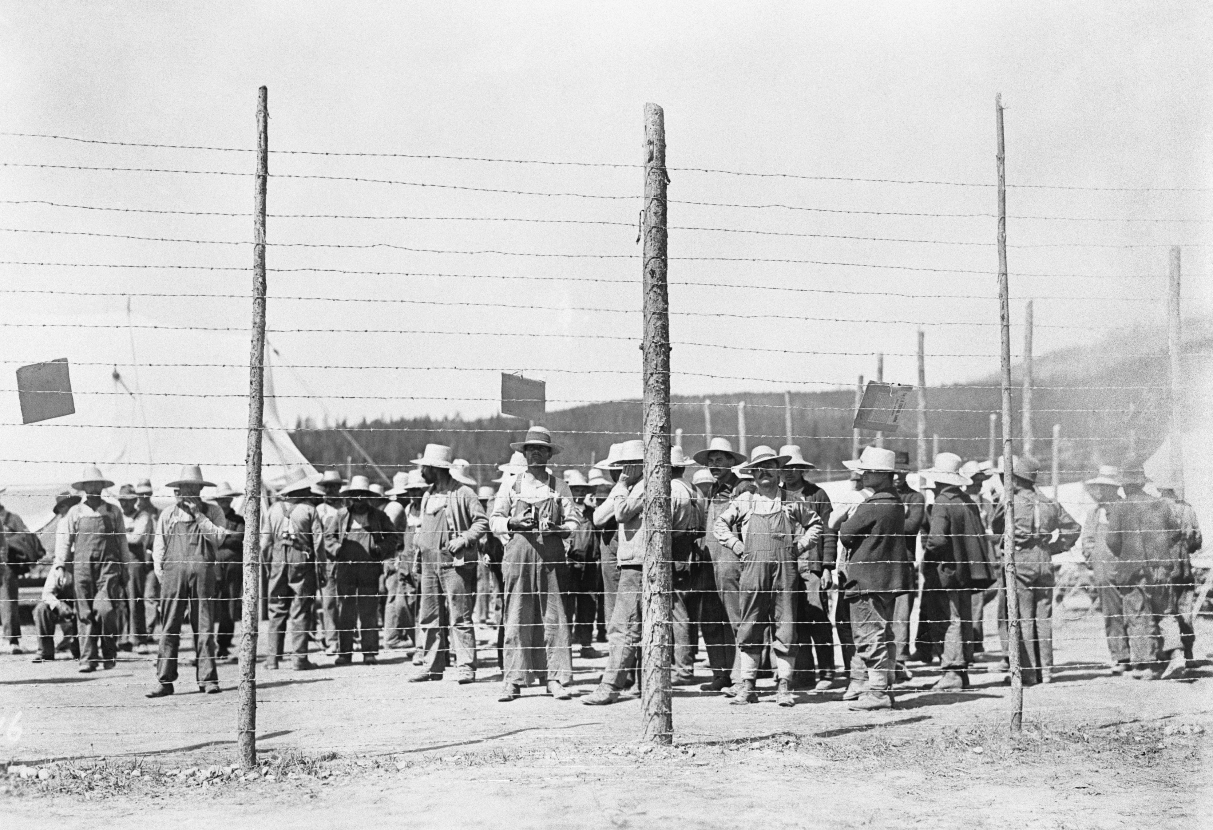 Photographie en noir et blanc d’hommes derrière une clôture de barbelés.//Black-and-white photograph of men behind a barbed-wire fence.