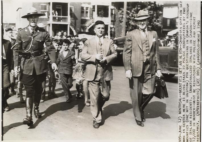 Un homme en costume est conduit par un agent de la GRC et un autre homme en complet avec un chapeau, portant une mallette.//A man in a suit is accompanied by an RCMP officer and another man in a suit and hat, carrying a briefcase.