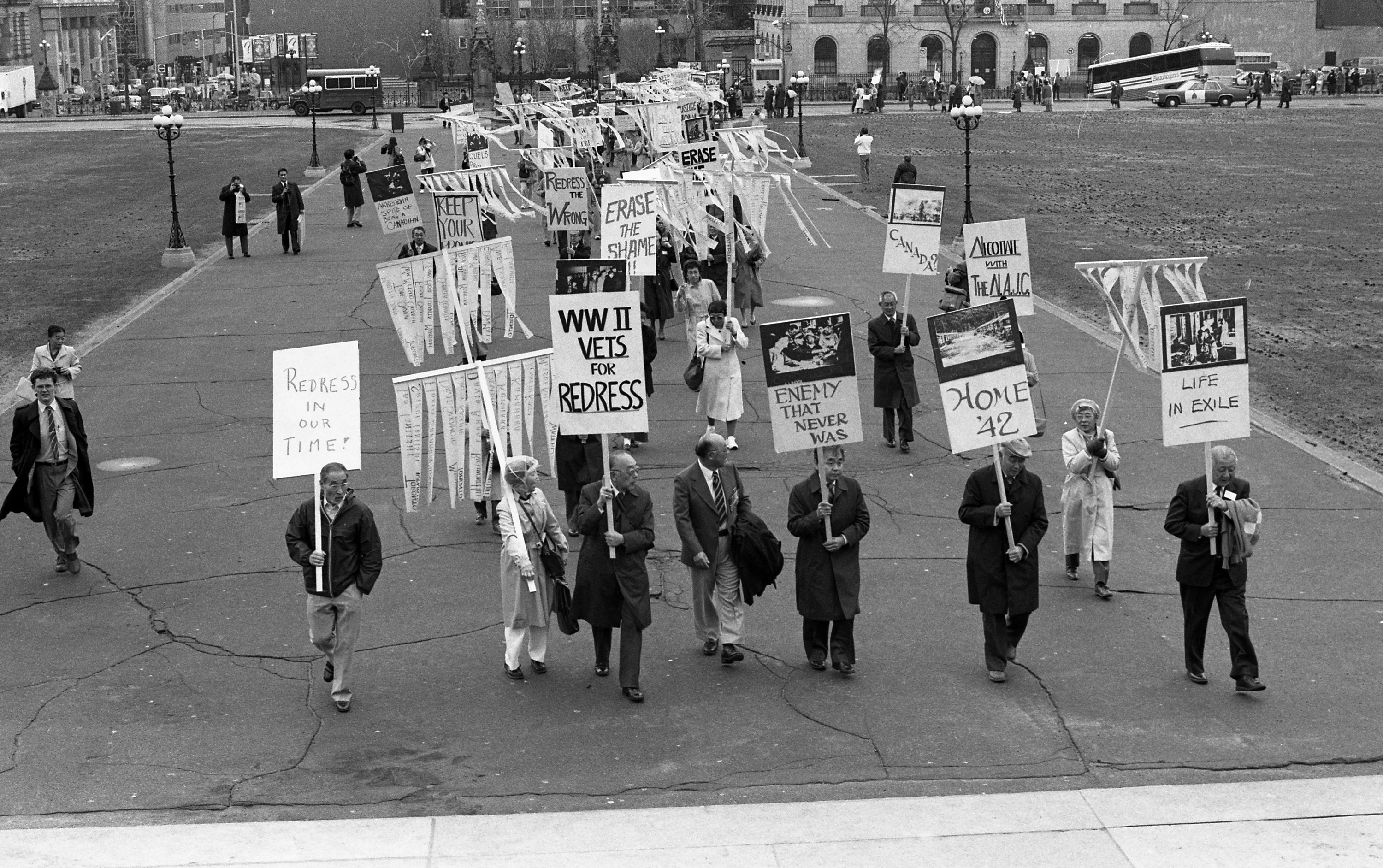 Photographie en noir et blanc de personnes marchant sur la colline du Parlement avec des pancartes. // Black-and-white photograph of people marching on Parliament Hill, holding signs.