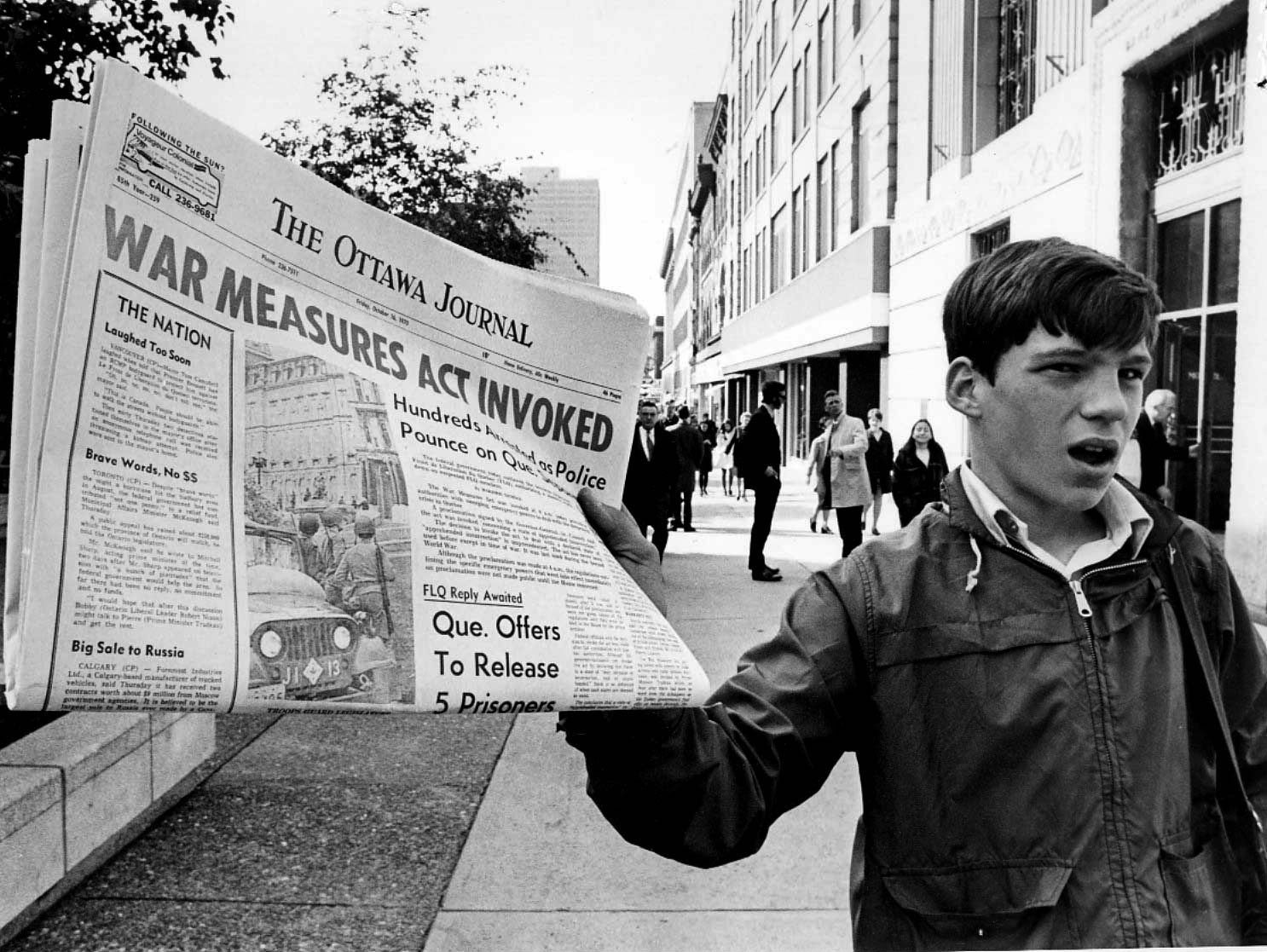 Photographie en noir et blanc d’un camelot tenant un journal // Black-and-white photograph of a paperboy holding a newspaper.