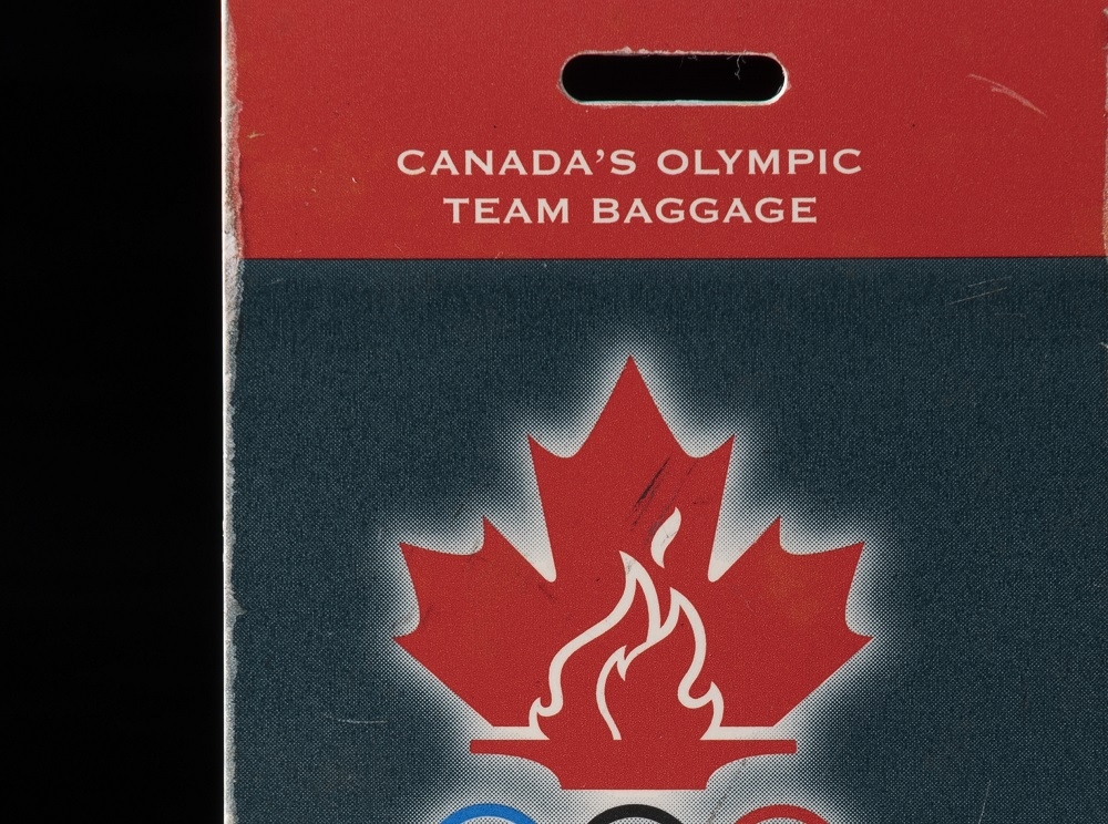 Étiquette rectangulaire bleue et rouge avec une feuille d’érable rouge et des anneaux olympiques.//Rectangular blue and red tag with a red maple leaf and Olympic rings