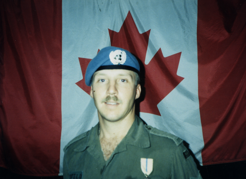 Man in peacekeeper beret in front of a Canadian flag