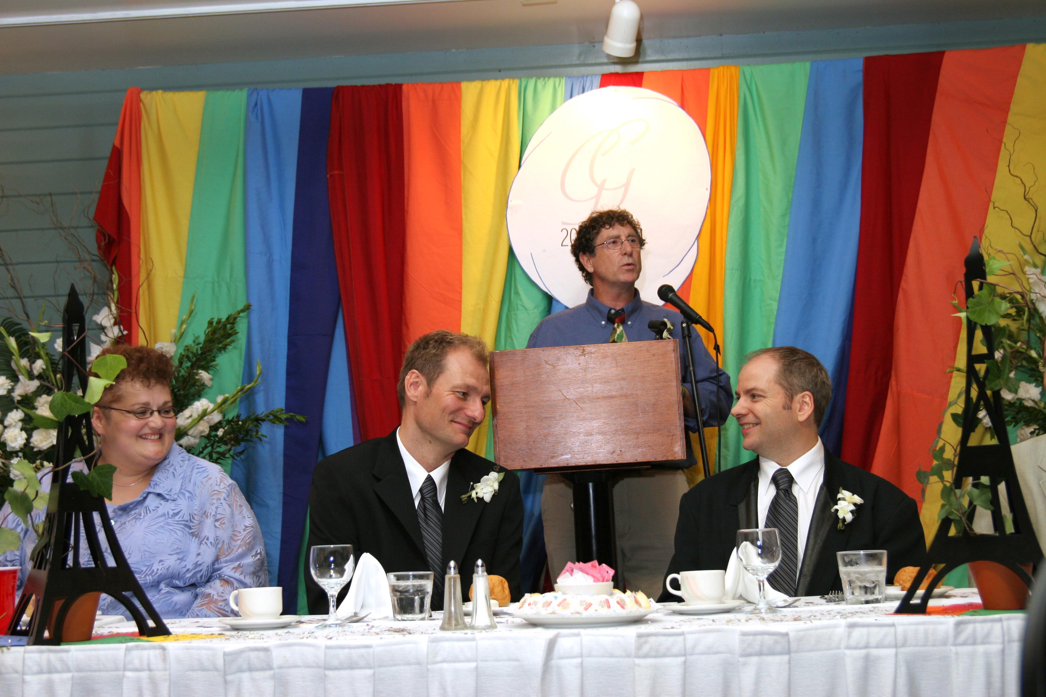 Deux hommes sont assis à une table d’honneur. Derrière eux, un homme parle sur un podium, devant une toile de fond arc-en-ciel.//Two men sit at a head table, a man speaks at a podium behind them in front of a rainbow backdrop