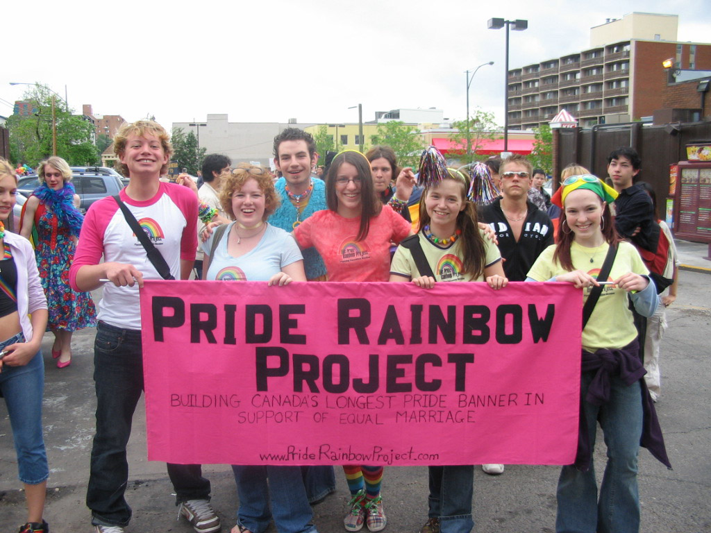Un groupe se tient derrière une bannière rose intitulée « Pride Rainbow Project ».//Group standing behind pink banner titled “Pride Rainbow Project”