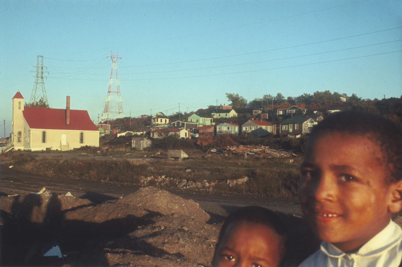 Colour photograph of two children in foreground, with white clapboard church in background. - Une photographie en couleur de deux enfants au premier plan avec une église à clins blancs à l’arrière-plan.