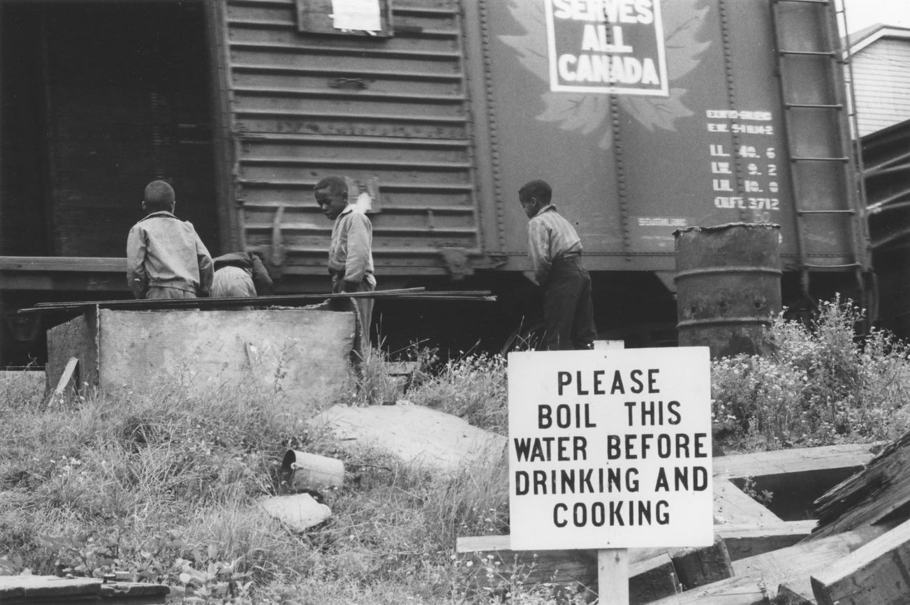 Black-and-white photograph of a group of children playing outside near a railway car, and a large sign with a boil-water advisory in the foreground. - Une photographie en noir et blanc d’un groupe d’enfants jouant dehors près d’un wagon et d’un grand panneau au premier plan précisant qu’il faut bouillir l’eau.