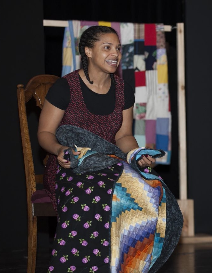 Colour photograph of a young Black woman sitting on a chair, with a colourful quilt on her lap. - Une photographie en couleur d’une jeune femme noire assise sur une chaise, avec une courtepointe colorée sur les genoux.