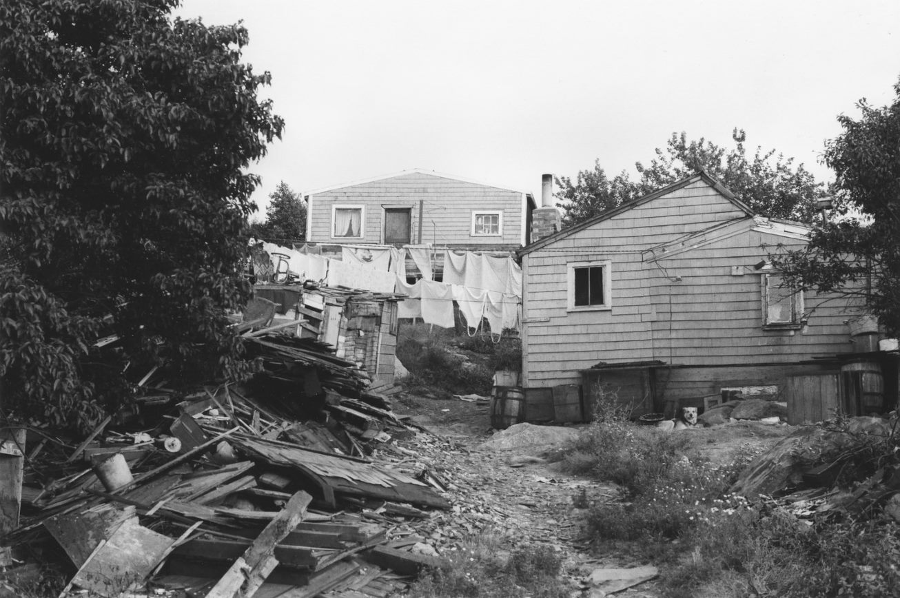 Black-and-white photograph of two houses, with a clothesline and laundry strung between them. - Une photographie en noir et blanc de deux maisons avec une corde à linge tendue entre elles et du linge.