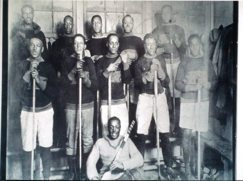 Black-and-white photograph of a group of men in old-fashioned hockey uniforms, holding hockey sticks. - 