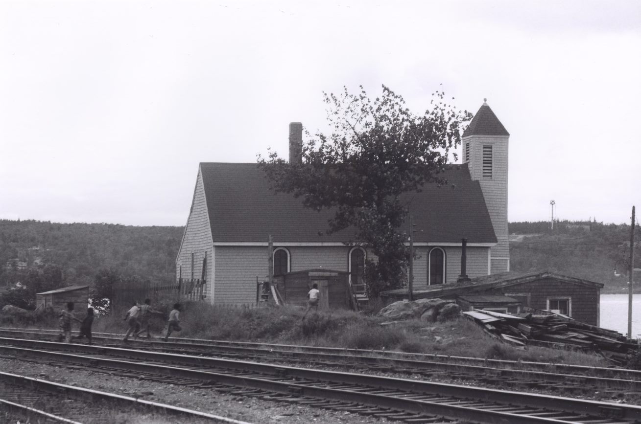 Black-and-white photograph of clapboard church with train tracks in foreground. - Une photographie en noir et blanc d’une église à clins avec une voie ferrée au premier plan.