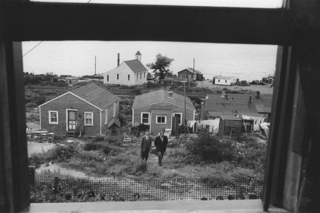 Black-and-white photograph of two city officials in suits, walking away from houses in the background. - Une photographie en noir et blanc de deux fonctionnaires municipaux en costume, s’éloignant de maisons à l’arrière-plan.