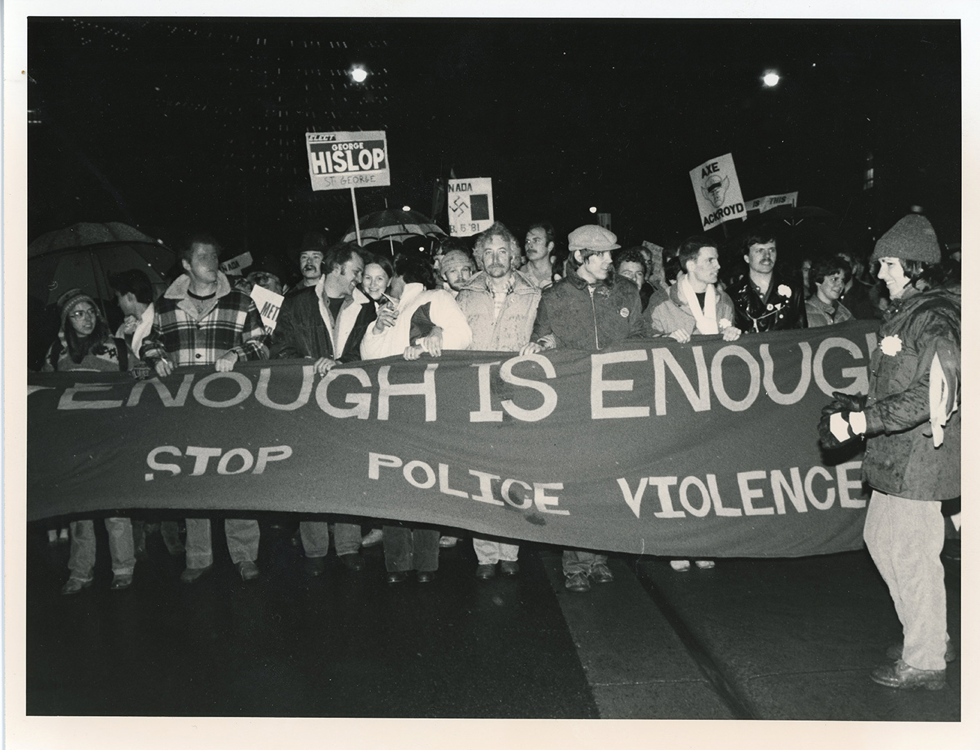 Photographie en noir et blanc d’hommes portant des pancartes, derrière une banderole sur laquelle on peut lire « Enough is Enough: Stop Police Violence ». Black-and-white photograph of men with signs, behind a banner that reads, “Enough is Enough: Stop Police Violence.”